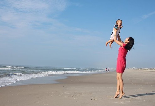 Frau mit hochgehaltenem Kind am Strand im Sommer.